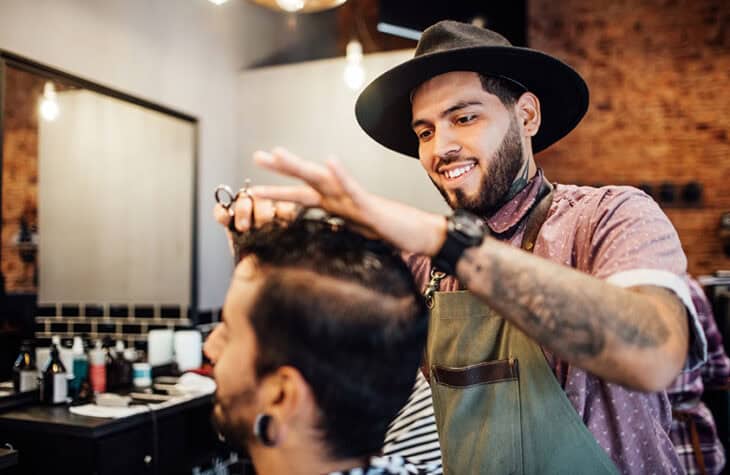 Smiling young hairdresser cutting customer's hair. Confident hipster barber is giving customer new hairstyle. They are at hair salon.