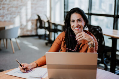 Business owner with calculator, spreadsheet, and laptop, looking into camera.