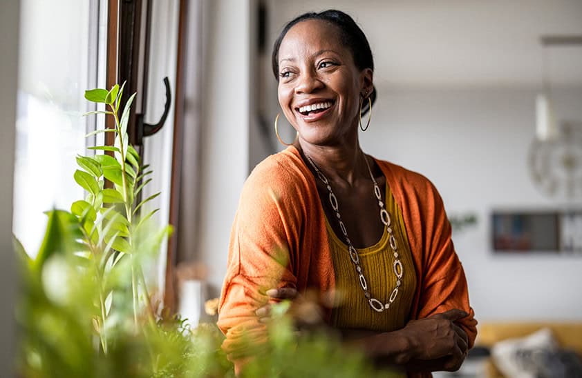 An older woman standing in their home and looking out the window with a smile.