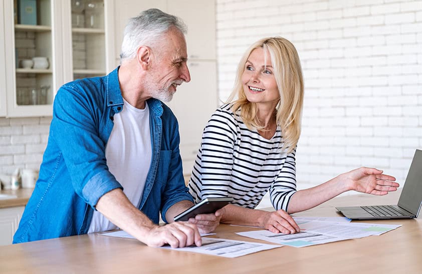 An older couple looking at each other while discussing documents viewed on a laptop.