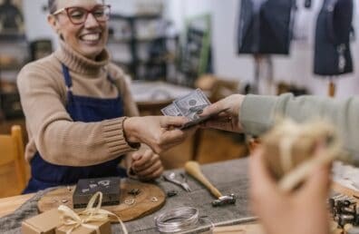 Close-up of two people exchanging a wrapped gift and cash over a table cluttered with tools and materials, highlighting a moment of transaction and exchange.