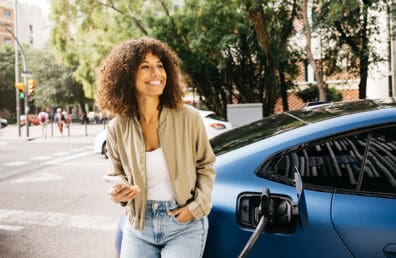 A woman holding her phone and leaning against her blue electric vehicle.