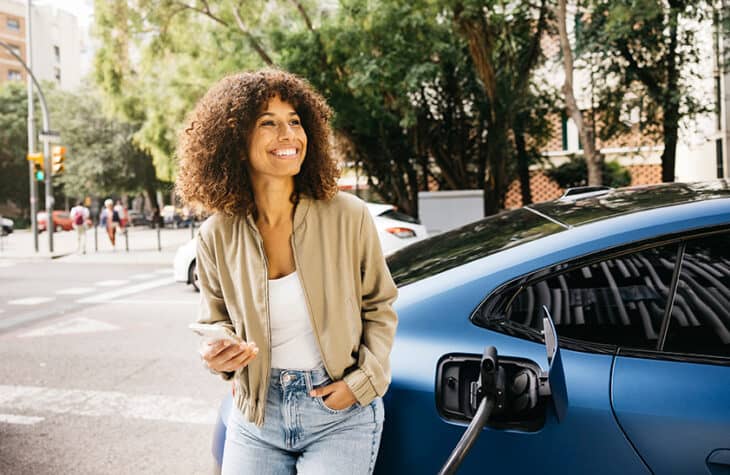 A woman holding her phone and leaning against her blue electric vehicle.