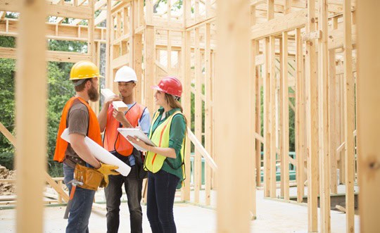 Latin descent female foreman or inspector checks blueprints in construction building site as other workers listen to instructions. Unfinished framed building in background.
