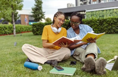 Two young female students reading books together and cheerfully discussing, while sitting on a grass area